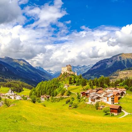 Schloss Tarasp auf einem Hügel, umgeben von grünen Wiesen und Dorfhäusern, mit Bergen und blauem Himmel im Hintergrund.