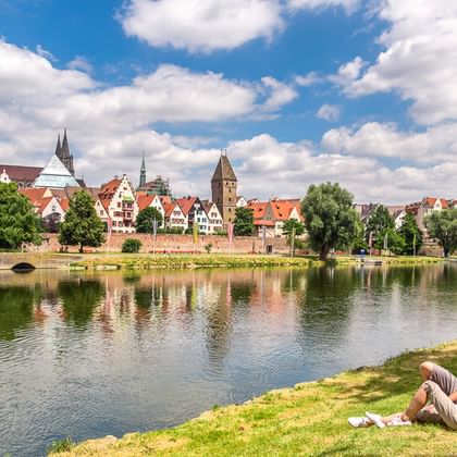 Zwei Radfahrer rasten am grasbewachsenen Donauufer mit Fahrrädern in der Nähe. Historische Stadt mit Kirchtürmen und bunten Gebäuden spiegelt sich im Wasser.