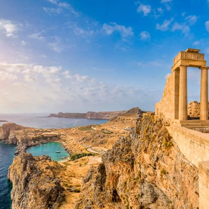 Antike griechische Säulen der Lindos Akropolis auf einer Klippe mit Blick auf das Mittelmeer und die felsige Küste mit kleiner Bucht.