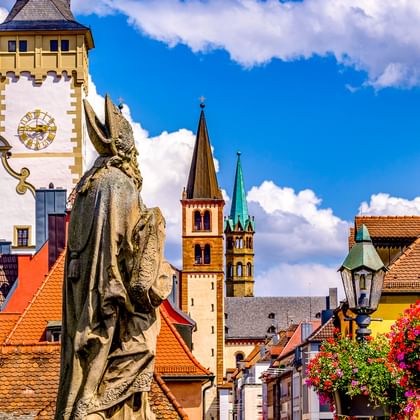 Steinstatue eines Heiligen auf der Alten Mainbrücke mit Würzburger Altstadtsilhouette, Kirchtürmen, Uhrturm und roten Ziegeldächern.