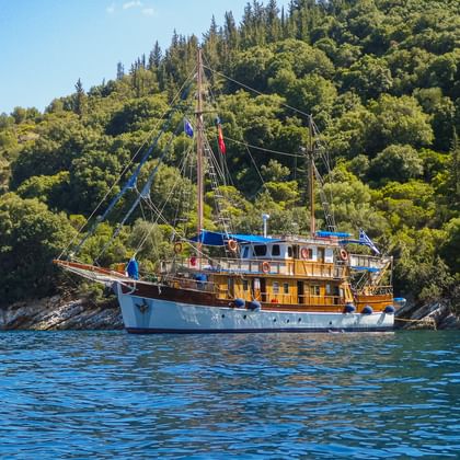 MS Panagiota, ein traditionelles Holzsegelschiff mit weiß-blauem Rumpf, vor Anker in ruhigem blauen Wasser nahe der bewaldeten griechischen Küste.