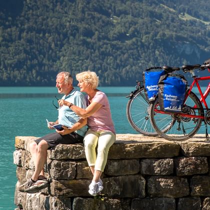 Zwei Radfahrer sitzen auf einer Steinmauer neben türkisfarbenem Schweizer See mit Tourenrädern. Bewaldete Berge im Hintergrund.