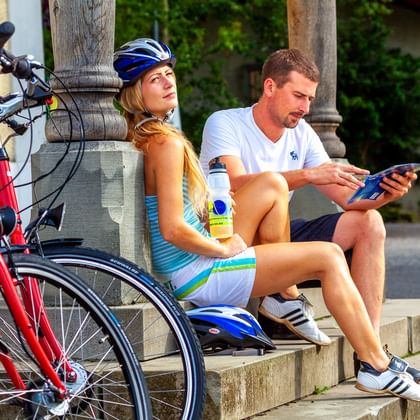 Zwei Radfahrer sitzen mit roten Fahrrädern auf Steinstufen und machen Pause. Frau mit blauem Helm hält Getränk, Mann prüft Karte am Bodensee.