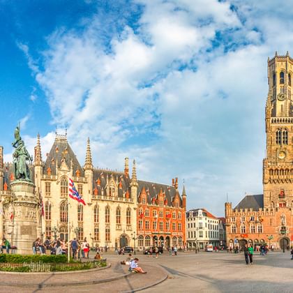 Brügger Marktplatz mit Belfried-Turm, historischen Gebäuden und Denkmal. Touristen versammeln sich auf dem Platz unter blauem Himmel.