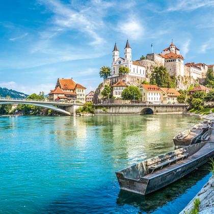 Historische europäische Stadt am türkisfarbenen Fluss mit Schloss, Kirchtürmen und Brücke. Holzboote am Steinufer unter blauem Himmel.