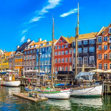 Historische Holzsegelboote am Nyhavn-Kanal in Kopenhagen mit bunten Stadthäusern aus dem 17. Jahrhundert unter blauem Himmel.