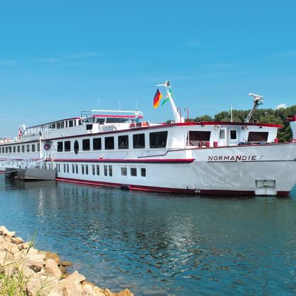 Weißes Flusskreuzfahrtschiff MS Normandie mit rotem Rand fährt auf blauem Wasser unter klarem Himmel, grüne Bäume und felsiges Ufer sichtbar.