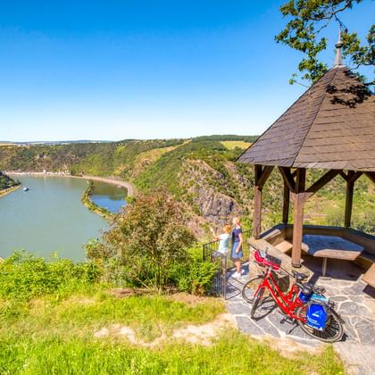 Wooden gazebo at Maria Ruh viewpoint with red bicycles, overlooking the Rhine River valley. Green hills and a curved river are visible below.