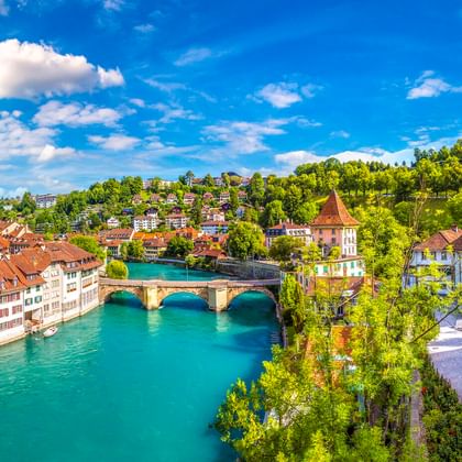 Nydeggbrücke über die türkisfarbene Aare in Bern mit historischen Gebäuden, roten Ziegeldächern, Kirchturm und grünem Hang unter blauem Himmel.