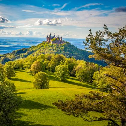 Burg Hohenzollern thront auf einem bewaldeten Hügel bei Hechingen, umgeben von grünen Wiesen und Bäumen unter blauem Himmel mit weißen Wolken.