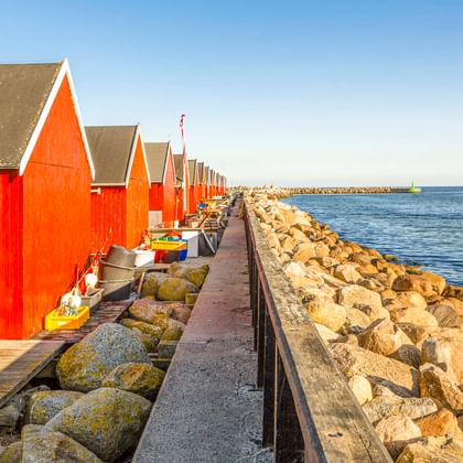 Reihe roter Holzbootshäuser mit weißer Verzierung entlang eines Steinpiers im Hafen von Rødvig. Kleine Boote neben den Gebäuden, ruhige blaue See.