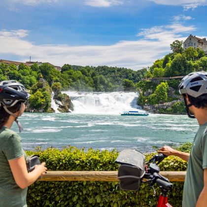 Zwei Radfahrer mit Helmen betrachten den Rheinfall von einer Aussichtsplattform. Ein Ausflugsboot fährt im türkisfarbenen Wasser unterhalb des breiten Wasserfalls.