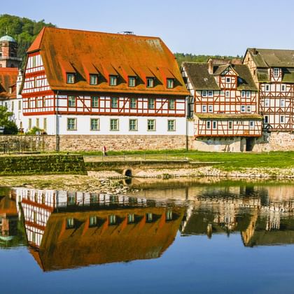 Traditionelle Fachwerkhäuser mit orangen Dächern entlang der Fulda in Rotenburg, die sich im ruhigen Wasser spiegeln, mit grünen Hügeln dahinter.