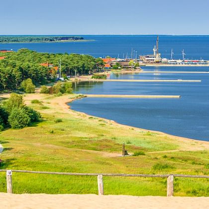 Panoramablick auf die litauische Küste mit grünen Wiesen, Sandstrand, blauem Buchwasser und Marina mit Segelbooten in der Ferne.