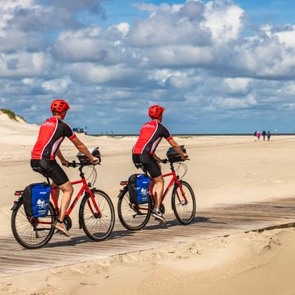 Zwei Radfahrer in roten Trikots und Helmen fahren am breiten Sandstrand von St. Peter-Ording mit Dünen und blauem Wolkenhimmel.