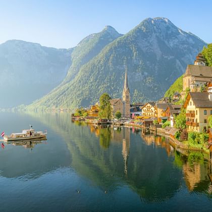 Hallstatt mit Kirchturm am ruhigen Seeufer, umgeben von bewaldeten Bergen. Ein Boot schwimmt auf dem spiegelnden Wasser.