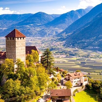 Mittelalterliche Burg mit Turm auf einem Hügel umgeben von grünen Bäumen, mit Blick auf das Etschtal und Berge in Südtirol unter blauem Himmel.