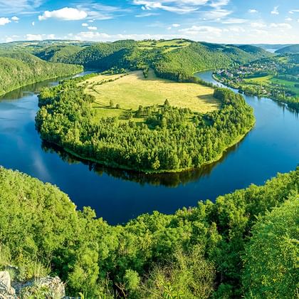 Luftaufnahme einer hufeisenförmigen Flussbiegung, umgeben von dichten Wäldern und grünen Wiesen unter blauem Himmel mit weißen Wolken in Tschechien.