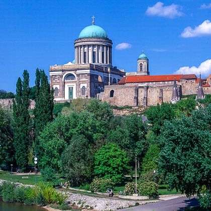 Die Basilika von Esztergom mit türkisfarbener Kuppel krönt einen Hügel über der Donau. Bäume säumen das Flussufer und ein Weg verläuft am Wasser.