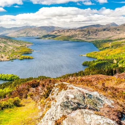 Panoramablick auf Loch Katrine, umgeben von grünen Wäldern und Hügeln. Felsiges Gelände im Vordergrund, blauer Himmel mit weißen Wolken.