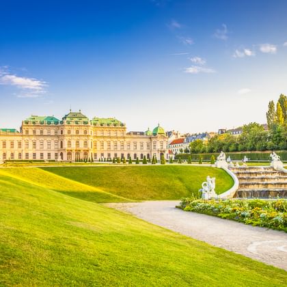 Schloss Belvedere in Wien mit symmetrischen Barockgärten, weißen Statuen, Brunnen und gepflegten Rasenflächen unter blauem Himmel.