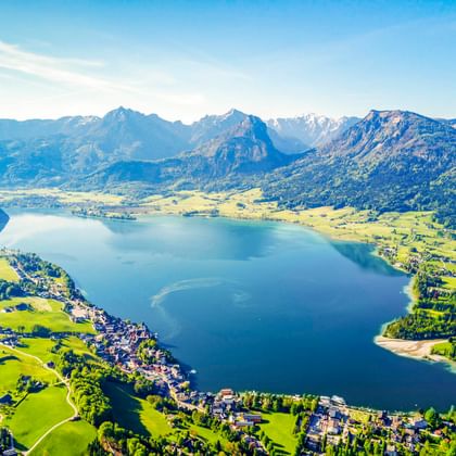 Aerial view of Wolfgangsee surrounded by green meadows and mountains. Small villages line the shore, with Alpine peaks in the background.