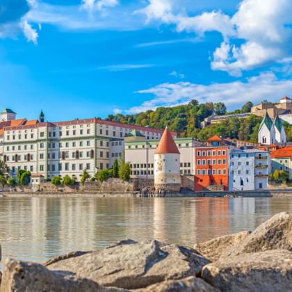 Bunte historische Gebäude der Passauer Altstadt an der Donau mit weißem Turm mit rotem Dach, vom felsigen Flussufer unter blauem Himmel.