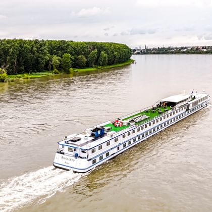 Luftaufnahme des Flusskreuzfahrtschiffs MS Rigoletto auf einem breiten Fluss. Grüne Wälder säumen das linke Ufer, eine Stadt ist in der Ferne sichtbar.