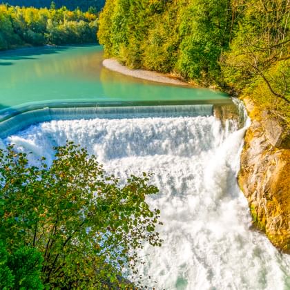 Luftaufnahme des Lechfalls bei Füssen mit türkisfarbenem Wasser, das über ein geschwungenes Wehr fließt, umgeben von dichtem grünem Wald und Felsen.