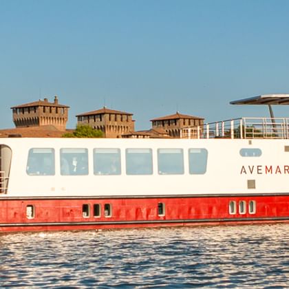 Flusskreuzfahrtschiff MS Ave Maria mit rotem Rumpf und weißem Aufbau auf ruhigem Wasser. Historische Gebäude im Hintergrund.