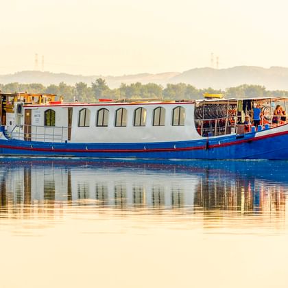 MS L'Estello, ein blau-weißes Flusskreuzfahrtschiff mit Bogenfenstern, gleitet auf ruhigem Wasser mit sichtbarer Spiegelung. Grünes Ufer im Hintergrund.