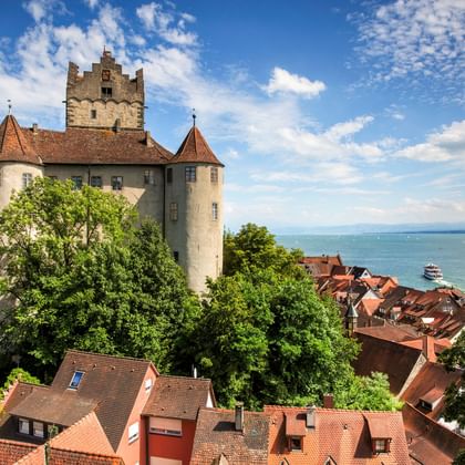 Burg Meersburg mit Turm und roten Ziegeldächern, umgeben von Bäumen, mit Blick auf den Bodensee mit Booten auf blauem Wasser unter bewölktem Himmel.