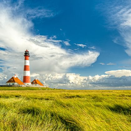 Rot-weiß gestreifter Leuchtturm Westerhever mit Nebengebäuden auf Westerheversand, umgeben von grünen Salzwiesen unter dramatischem Wolkenhimmel.
