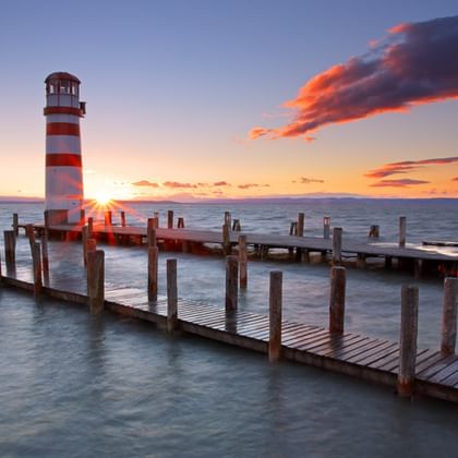 Rot-weiß gestreifter Leuchtturm auf Holzsteg am Neusiedler See bei Sonnenuntergang mit orangefarbenen Wolken und ruhigem Wasser.