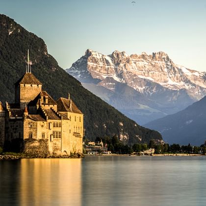 Schloss Chillon am Genfersee mit schneebedeckten Bergen im Hintergrund. Die mittelalterliche Festung steht im goldenen Licht am Wasser.