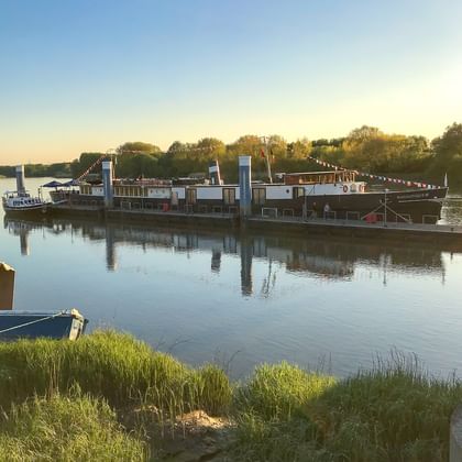Flusskreuzfahrtschiff MS Magnifique III bei Sonnenuntergang mit bunten Flaggen vertäut. Bäume säumen das gegenüberliegende Ufer unter klarem Himmel.