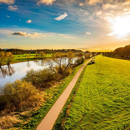 Asphaltierter Radweg entlang der Weser bei Porta Westfalica bei Sonnenuntergang. Grüne Wiesen säumen den Weg, Bäume stehen im Wasser, Hügel im Hintergrund.
