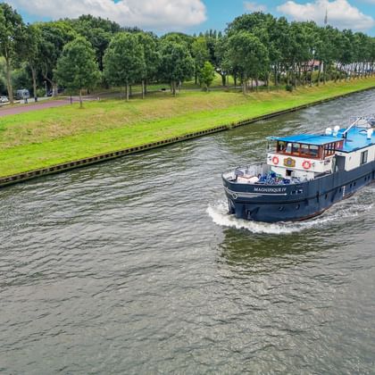 MS Magnifique III river cruise ship sailing on a Belgian canal lined with green trees. The blue and white vessel moves through calm water.