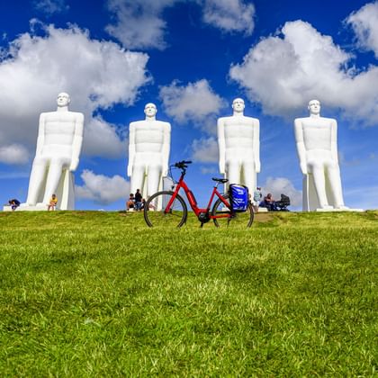 Red touring bicycle with blue panniers on green grass in front of four large white human statues against a blue sky with white clouds.