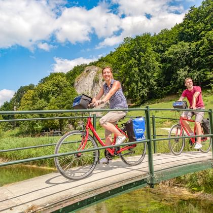 Zwei Radfahrer auf einer Metallbrücke über einen Bach bei Schelklingen. Grüne bewaldete Hügel und Felsformationen im Hintergrund unter blauem Himmel.