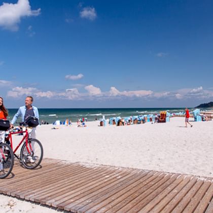 Zwei Radfahrer mit roten Rädern auf Holzsteg am Ostseestrand mit weißem Sand, blauen Strandkörben und Menschen an einem sonnigen Tag.