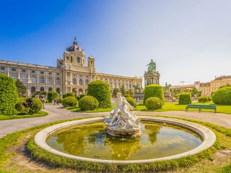 Circular fountain with white sculpture in manicured park. Kunsthistorisches Museum Vienna with dome and equestrian statue visible behind.
