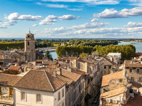 Luftaufnahme der Altstadt von Arles mit Terrakotta-Dächern, einem Kirchturm aus Stein und der Rhône mit grüner Vegetation unter blauem Himmel.
