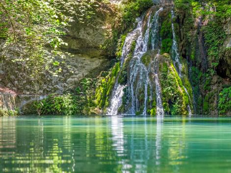 Wasserfall, der über moosbewachsene Felsen in ein türkisfarbenes Becken im Petaloudes-Tal, Rhodos, fällt, umgeben von üppiger Vegetation.