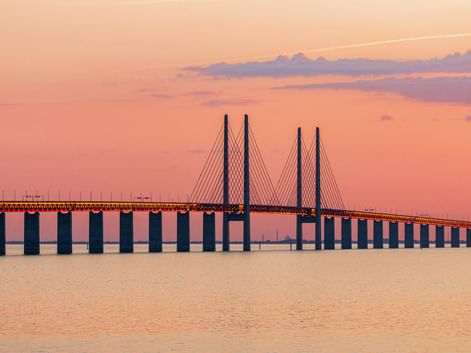 Die Öresundbrücke erstreckt sich über ruhiges Wasser unter einem rosa-orangefarbenen Sonnenuntergangshimmel. Die Schrägseilbrücke mit hohen Pylonen.
