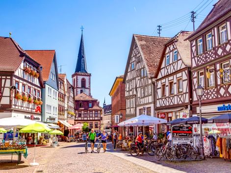 Fußgängerzone mit traditionellen Fachwerkhäusern, Kirchturm, Straßencafés und Radfahrern. Bunte Gebäude säumen Kopfsteinpflasterstraße.