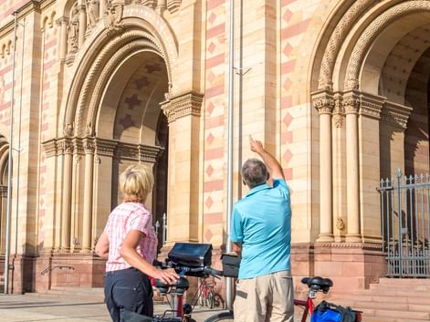Zwei Radfahrer mit Tourenrädern und blauen Taschen stehen vor der gestreiften romanischen Fassade des Speyerer Doms mit verzierten Portalen.