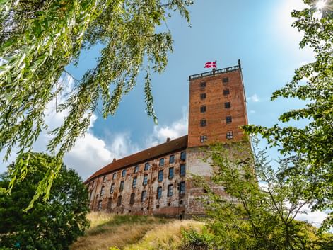 Koldinghus Schloss in Kolding mit rotem Backsteinturm und dänischer Flagge, eingerahmt von grünen Bäumen unter blauem Himmel mit weißen Wolken.