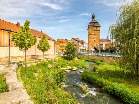 Bach fließt durch Bad Staffelstein Innenstadt mit historischem Steinturm, bunten Häusern und orangefarbenen Ziegeldächern unter blauem Himmel.