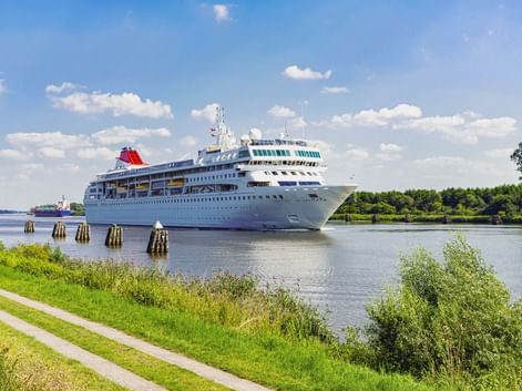 Weißes Kreuzfahrtschiff mit rotem Schornstein auf dem Nord-Ostsee-Kanal. Grüne Ufer mit Radweg und Holzpfosten säumen die Wasserstraße unter blauem Himmel.